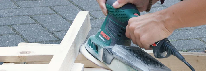 Person schleift Holzlatten mit einem Schwingschleifer.